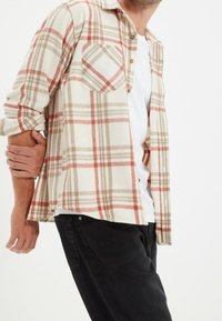 Camisa de cuadros beige con acentos en rojo y marrón, con cierre de botones en la parte delantera y un bolsillo en el pecho. Llevada sobre una camiseta blanca y pantalones negros.