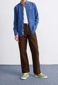 Hombre con camisa azul abotonada sobre camiseta blanca sin mangas, pantalones marrones y zapatillas verde claro, de pie sobre un suelo con alfombra azul.