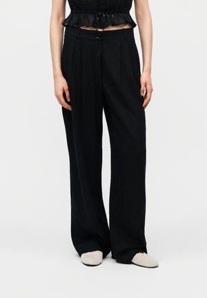 Kobieta w czarnych szerokich spodniach, czarnym, krótkim, falbankowym topie oraz białych wsuwanych butach, stojąca na tle jednolitego jasnego tła.