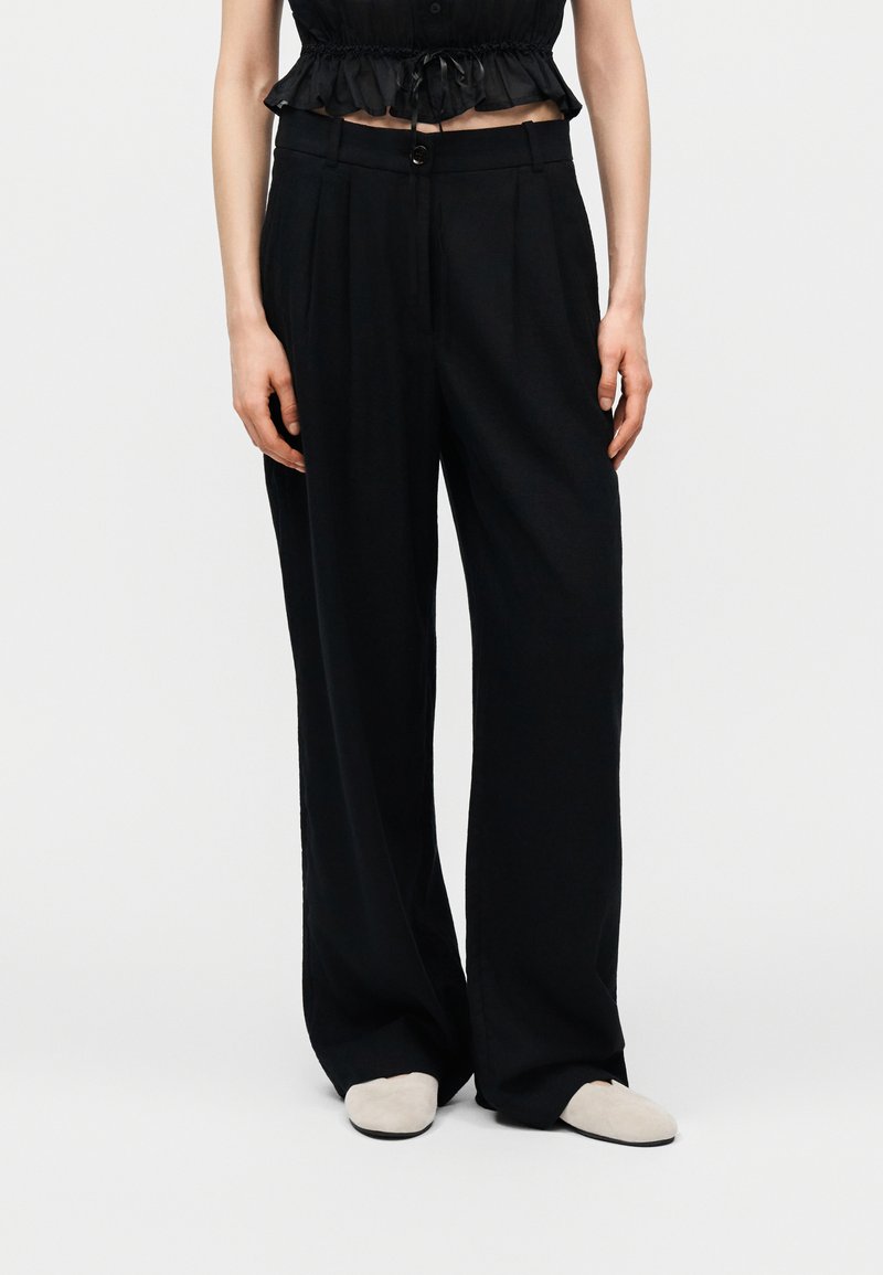 Γυναίκα που φοράει μαύρο παντελόνι με φαρδιά μπατζάκια, μαύρο κροπ τοπ με βολάν και λευκά παπούτσια χωρίς κορδόνια, στέκεται μπροστά σε ένα απλό φωτεινό φόντο.