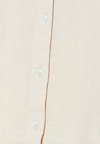 Camisa beige de botones con tejido texturizado. Presenta una fila de botones blancos en la parte delantera, con costuras visibles a lo largo de la tapeta.
