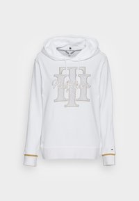 Felpa bianca con una grafica frontale realizzata in strass che presenta la scritta "Hilfiger" e un grande logo. Ha un cappuccio con cordino e dettagli dorati sui polsini.