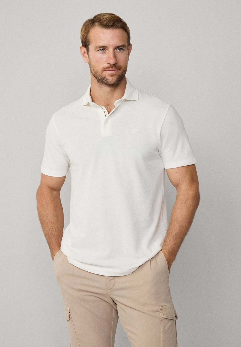 Polo bianco in cotone con maniche corte e colletto. Presenta una pattina decorativa a righe e un logo discreto sul petto.