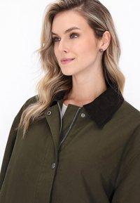 Femeie cu păr ondulat blond, îmbrăcată într-o jachetă verde măslinie cu guler închis, privind ușor spre dreapta pe un fundal simplu.