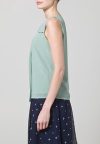Top sin mangas de color verde menta con un cuello redondo, hecho de tela suave, que presenta una abertura lateral, combinado con una falda oscura con pequeños patrones de pájaros.