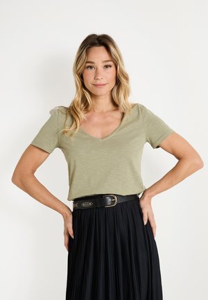 T-shirt à manches courtes de couleur vert olive clair avec un design à col en V, associé à une jupe plissée noire et une ceinture noire texturée.