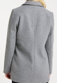 Manteau en laine grise à coupe ajustée, doté d'un col cranté, d'une texture lisse et d'un détail de couture au dos. Aucune fermeture ou ornement visible.