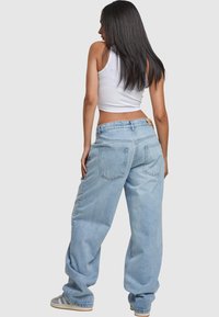 Woman with long dark hair wearing a white crop top, loose light blue jeans, and gray sneakers, standing with back turned on a plain background.