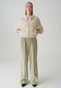 Hellbeige, flauschige Bomberjacke mit Reißverschluss, getragen über einem cremfarbenen Oberteil und kombiniert mit hoch taillierten, hellgrünen Cordhose und schwarzen High Heels.