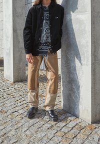 Sort fløjljakke med knapper, mønstret skjorte og lysebrune patchwork-bukser. Afsluttet med sorte læderstøvler. Står op ad en betonvæg.