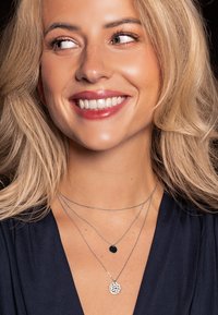 Collane stratificate in argento con un pendente rotondo texturizzato e un piccolo charm circolare nero, appese a catene sottili e indossate su una parte superiore scura.