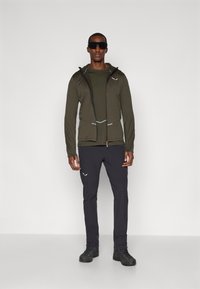 Ελιά πράσινη ζακέτα με φερμουάρ, μαύροι στενοί παντελόνες και μαύρα παπούτσια. Ανακλαστικές λεπτομέρειες στη ζακέτα; ορατό υφή υφάσματος. Μινιμαλιστικός σχεδιασμός.