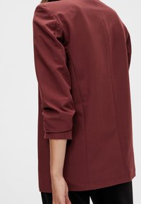 Personne portant un blazer rouge foncé à manches longues retroussées, vue de dos sur un fond clair uni.