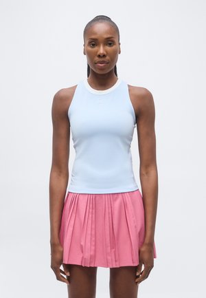 Kvinde stående med fronten mod kameraet iført en lyseblå ærmeløs top og en lyserød plisseret nederdel mod en ensfarvet baggrund.