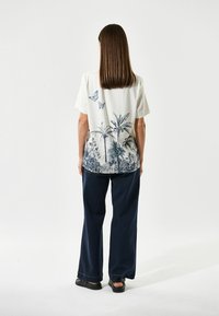 Hvid kortærmet bluse med et blåt botanisk print på ryggen, parret med brede mørkeblå bukser og sorte sandaler.