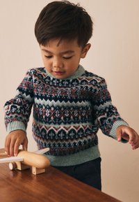 Stickad tröja med flerfärgade geometriska mönster i marinblått, grönt, rött och krämvitt. Barn leker med en träleksaksflygplan på ett bord.