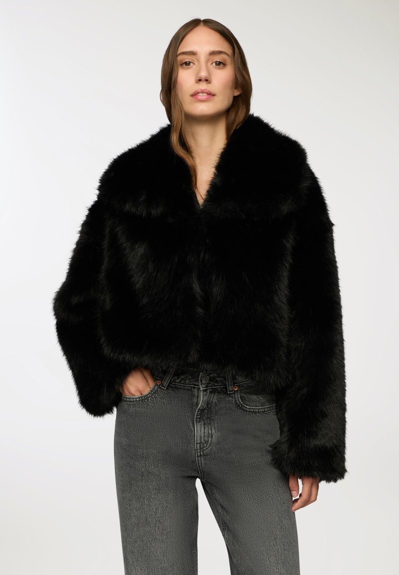 Czarna kurtka z sztucznego futra z krótkim krojem, szerokim kołnierzem i luźnymi rękawami. Połączona z szarymi dżinsami, które mają widoczne kieszenie i subtelną teksturę.