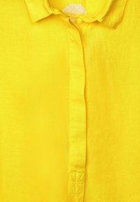 Camisa de lino amarilla con diseño de botones, cuello y tela texturizada con un acento cosido cerca de la abertura de los botones.