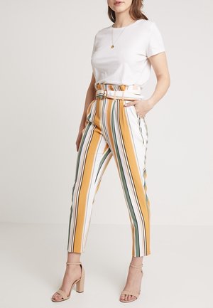 Nainen pukeutuneena korkeavyötäröisiin housuihin, joissa on keltaisia, vihreitä, valkoisia ja mustia pystysuoria raitoja ja vyö, valkoinen t-paita ja beigekorkoiset sandaletit.