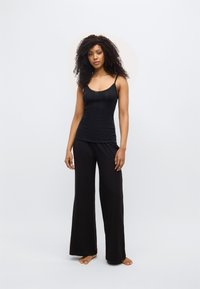 Μαύρο τοπ με λεπτές τιράντες, υφασμένο με υφή, σε συνδυασμό με φαρδιά μαύρη παντελόνα. Το μοντέλο στέκεται ξυπόλητο μπροστά σε φωτεινό φόντο.