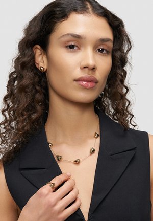 Frau mit lockigem Haar trägt abgestimmten goldfarbenen Ohrhänger, Halskette und Ring in Herzform, bekleidet mit einem ärmellosen schwarzen Oberteil mit Kragen.