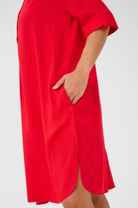 Robe rouge, de longueur genou, avec des manches courtes roulées, comportant une poche latérale et un tissu légèrement texturé. Des fentes latérales améliorent le confort et le mouvement.