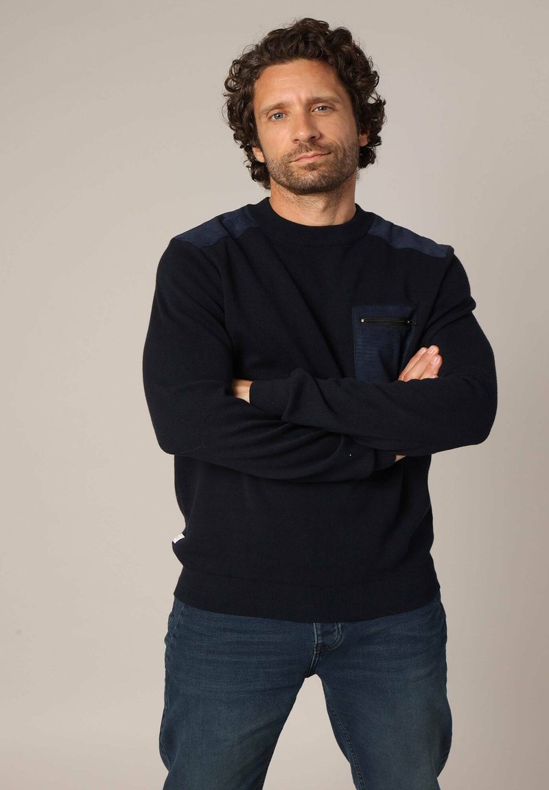 Pull en tricot bleu marine avec une texture lisse, doté de panneaux d'épaule bleu foncé et d'une poche poitrine zippée. Porté avec un jean bleu.