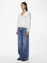 Bluză albă cu guler volan, margine din dantelă și închidere cu șnur. Asortată cu blugi cargo largi din denim albastru și pantofi negri.
