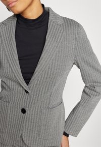 Femme portant un blazer gris à chevrons avec deux boutons noirs sur un col roulé noir, debout avec une main sur la hanche.