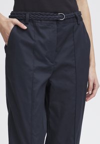 Pantaloni blu navy con cintura intrecciata con fibbia ovale in metallo; realizzati in tessuto liscio con tasche frontali e vestibilità rilassata.
