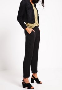 Chaqueta corta de mezclilla negra sobre una camiseta de color oliva claro. Pantalones negros entallados con el dobladillo enrollado. Sandalias negras de tacón bloque con detalle de hebilla.