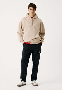 Sudadera beige con el texto "RED BULL RACING", superpuesta sobre una camiseta roja. Pantalones cargo negros con bolsillos, zapatillas blancas con suelas marrones.