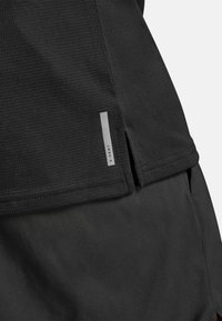 Černý atletický top z texturované látky s bočním rozparkem, s reflexní značkou na lemu. Jednoduchý design zaměřený na funkčnost.