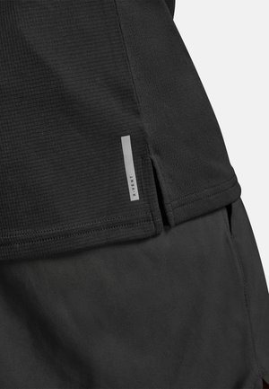 Musta urheilutoppi, valmistettu teksturoidusta kankaasta, jossa sivuhalkio ja heijastava merkki helmassa. Yksinkertainen muotoilu, joka keskittyy toiminnallisuuteen.