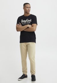 Schwarzes Baumwoll-T-Shirt mit dem Aufdruck "Stay Cool Blend Denim" in Weiß, kombiniert mit beigen Jogginghosen und schwarzen Sneakers, Arme verschränkt.