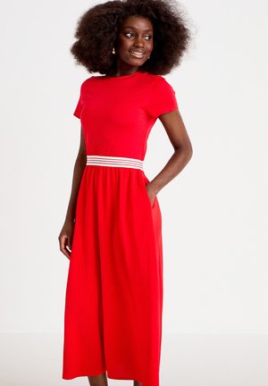 Kvinde med voluminøst krøllet hår iført en lys rød midi-kjole med korte ærmer og hvidstribet taljebånd, hænderne i lommerne, smilende og kiggende mod højre.