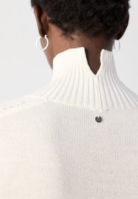 Gros plan sur le cou et le haut du dos d'une personne portant un pull à col roulé côtelé crème avec une petite étiquette de marque en métal sous le col.