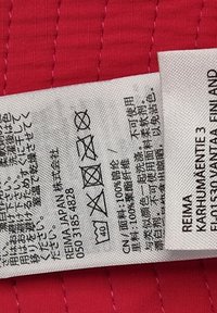 Czerwone tło z tkaniny z białą metką wyprodukcji, na której znajdują się symbole i tekst. Metka zawiera instrukcje prania oraz informacje o materiale.