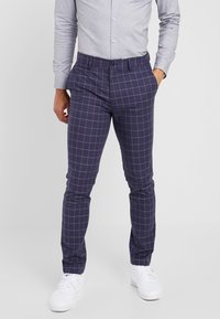 Man som har på sig marinblå glest rutiga byxor med vita rutlinjer, en ljusgrå knappad skjorta och vita sneakers, står mot en enfärgad bakgrund.