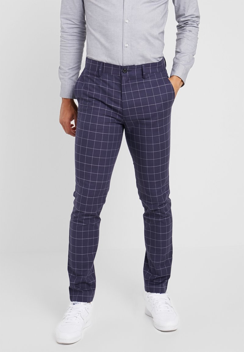 Man som har på sig marinblå glest rutiga byxor med vita rutlinjer, en ljusgrå knappad skjorta och vita sneakers, står mot en enfärgad bakgrund.