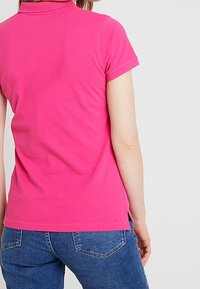 Helles pinkes Kurzarm-Poloshirt mit kragenartigem Ausschnitt, weicher Textur und seitlichen Schlitzen, kombiniert mit blauen Jeans.