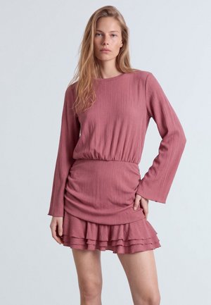 Donna con lunghi capelli biondi che indossa un abito rosa polvere a maniche lunghe, con gonna arricciata e orlo a balze sovrapposte.