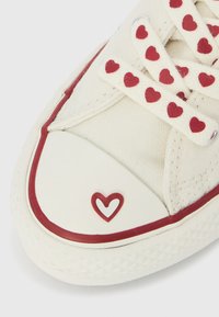 Πάνινο αθλητικό παπούτσι σε κρεμ χρώμα με κορδόνια με κόκκινα μοτίβα καρδιών, λευκό καπάκι από καουτσούκ στη μύτη και κόκκινη διακοσμητική λεπτομέρεια. Διαθέτει λογότυπο καρδιάς στη μύτη.