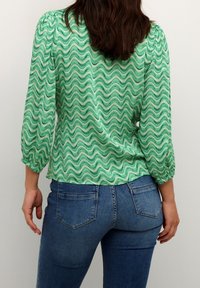 Una donna che indossa jeans blu e una blusa verde con un motivo a linee ondulate, in piedi di spalle contro uno sfondo chiaro e uniforme.