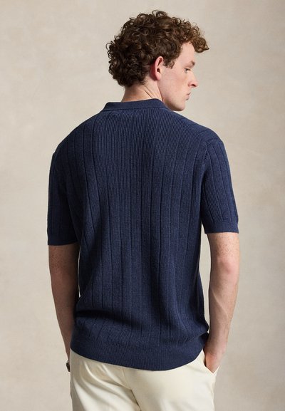 Homme aux cheveux bouclés portant une chemise à manches courtes côtelée bleu marine et un pantalon beige clair, debout les mains dans les poches, tourné légèrement de côté et en arrière.