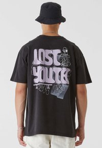 T-shirt en coton noir avec un grand texte graphique rose "LOST YOUTH" au dos, présentant des illustrations stylisées et une coupe décontractée.