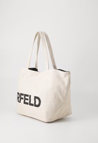 Beige Canvas-Shopper-Tasche mit schwarzem "CARLFELD"-Text. Verfügt über zwei lange Tragegriffe, einen offenen oberen Bereich und eine weiche Textur. Einfache, quadratische Form.