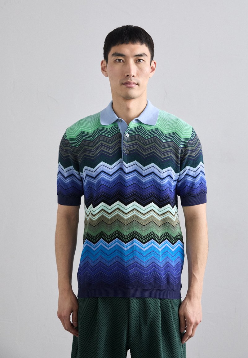 Polo tricotat în model zigzag verde, albastru și bleumarin, cu mâneci scurte și guler bleumarin deschis; trei nasturi în față.