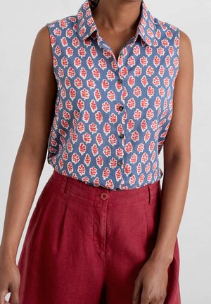 Mouwloze blauwe blouse met rood-witte bladerenpatronen, gestopt in een rood hooggetailleerde broek met knoopsluiting.