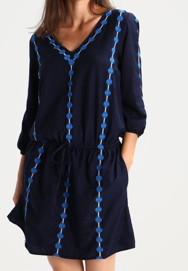 Femme portant une robe bleu marine à manches trois-quarts avec des lignes d'embroiement verticales bleues, cintrée à la taille par un cordon.